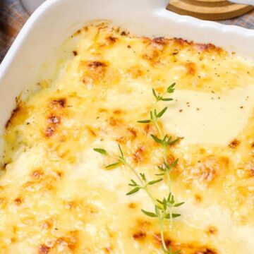 A close-up of a baked dish in a white rectangular casserole dish. The dish features a golden-brown, bubbling cheese topping with a sprig of fresh thyme placed diagonally in the center. A wooden pepper grinder is partially visible on the left.
