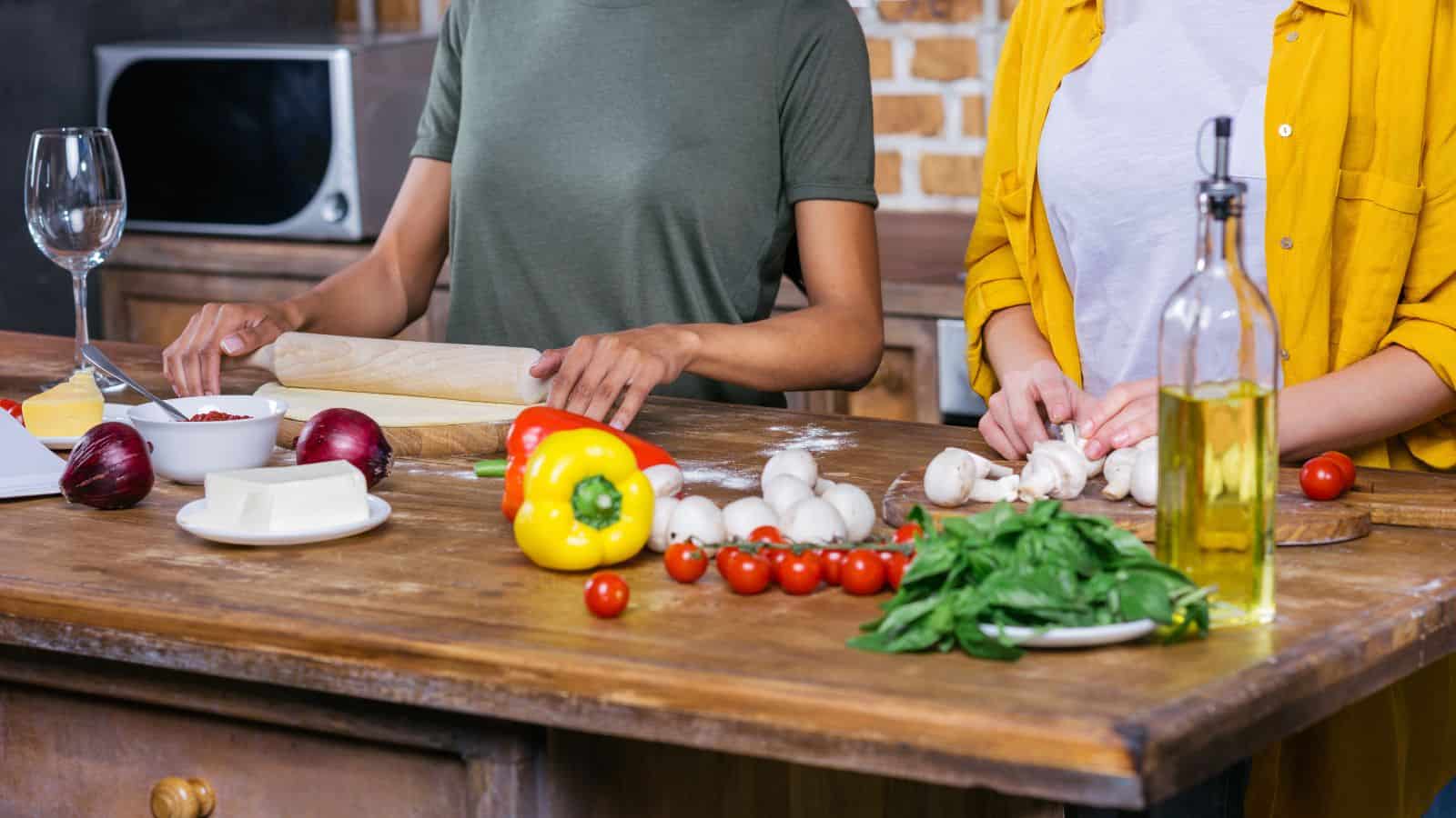Two people prepare food in a kitchen, seamlessly integrating holiday meal planning tips. One rolls dough with a rolling pin, while the other cuts mushrooms. Nearby are ingredients including bell peppers, cherry tomatoes, and fresh herbs. A bottle of olive oil and a microwave complete the cozy scene.