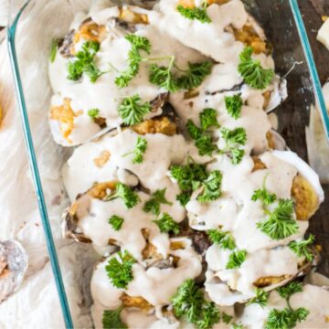 A glass dish filled with baked oysters topped with creamy sauce and garnished with parsley. Surrounding the dish are lemon halves, parsley sprigs, oyster shells, and garlic cloves on a wooden surface.