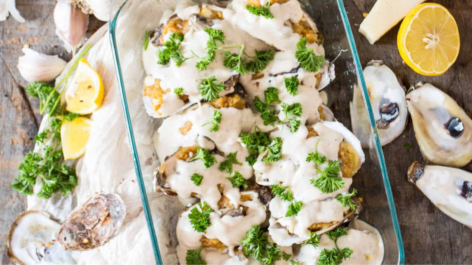 A glass dish filled with baked oysters topped with creamy sauce and garnished with fresh parsley. Lemon slices and oyster shells are placed nearby on a wooden surface, along with garlic cloves.