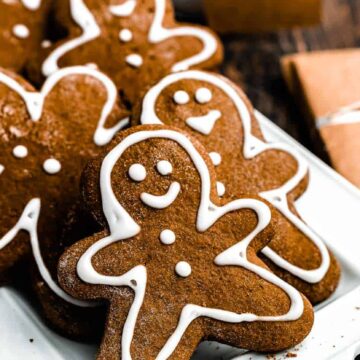A white tray of gingerbread cookies shaped like people, decorated with white icing for faces and outlines. A wrapped gift with a pine decoration is in the background on a dark wooden table.