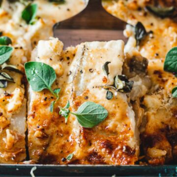 A baked dish featuring cheesy, golden-brown lasagna with visible layers and sprinkled with fresh herbs. Some pieces have been served, revealing a creamy interior.