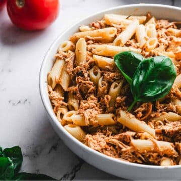 A bowl of penne pasta mixed with shredded meat and garnished with a basil leaf on a marble surface. Two tomatoes and a few basil leaves are nearby. A fork is placed in the bowl.