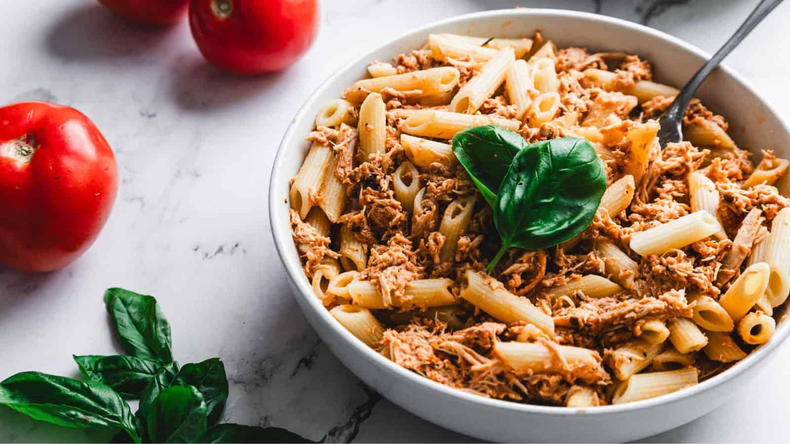 A bowl of penne pasta topped with shredded meat and garnished with fresh basil leaves. A fork is placed in the bowl. Red tomatoes and additional fresh basil leaves are on the marble surface nearby.