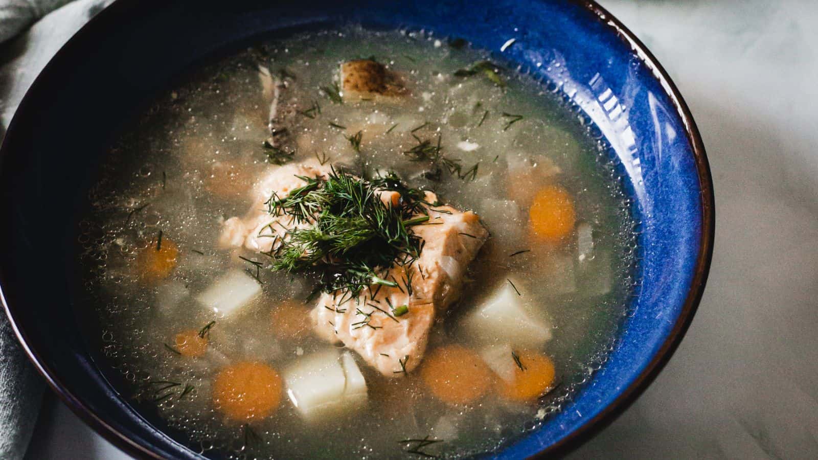 A blue bowl filled with fish soup containing chunks of fish, carrots, and potatoes, garnished with fresh dill. The soup has a clear broth with visible herbs.