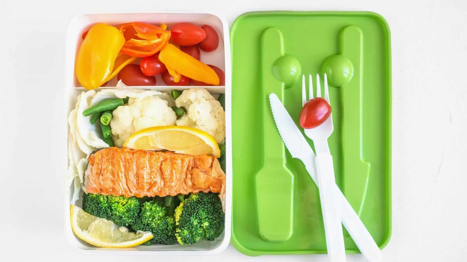 A bento box with compartments containing cherry tomatoes, sliced bell peppers, cauliflower, green beans, broccoli, and a piece of cooked salmon with lemon slices. A green lid holds a white fork, knife, spoon, and a single tomato.