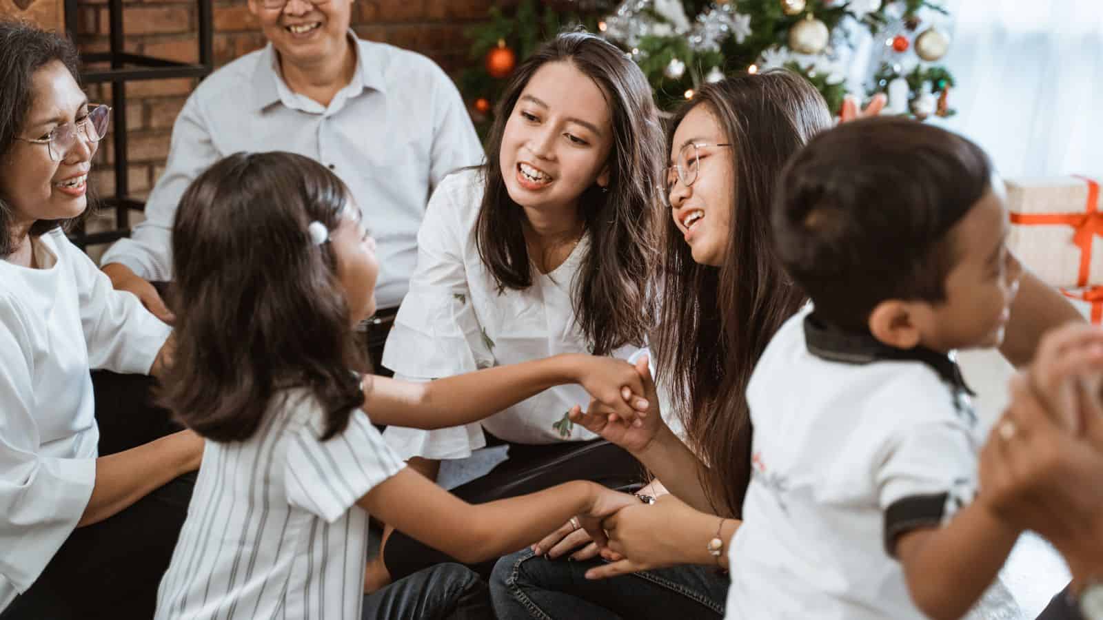 A group of people, including children and adults, sit together indoors, smiling and talking. A Christmas tree with ornaments and lights is visible in the background. The setting appears cozy and festive, creating a perfect atmosphere for a holiday feast.