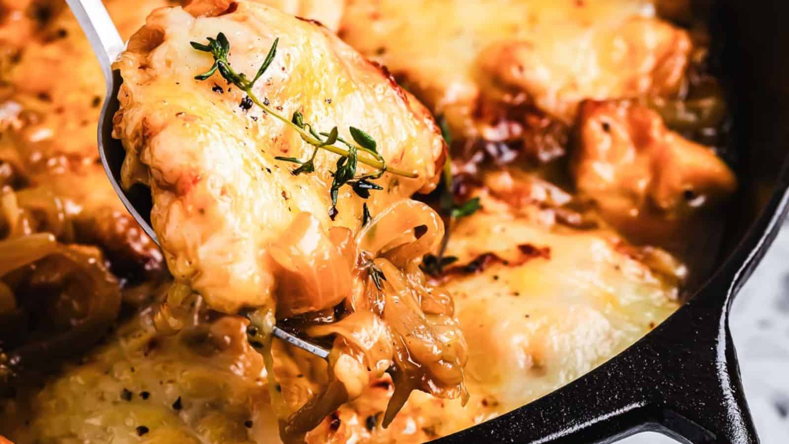 A close-up of a baked dish in a skillet featuring melted cheese, onions, and herbs. A serving spoon is lifting a portion, highlighting the golden-brown cheese and caramelized onions on top.