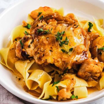 A white bowl filled with wide pasta noodles topped with cooked chicken in a creamy sauce, garnished with fresh herbs. A fork rests on a napkin beside the bowl.