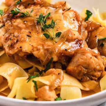 A plate of pappardelle pasta topped with tender chicken pieces and caramelized onions, garnished with fresh herbs. The sauce appears creamy, and the dish is served in a white bowl.