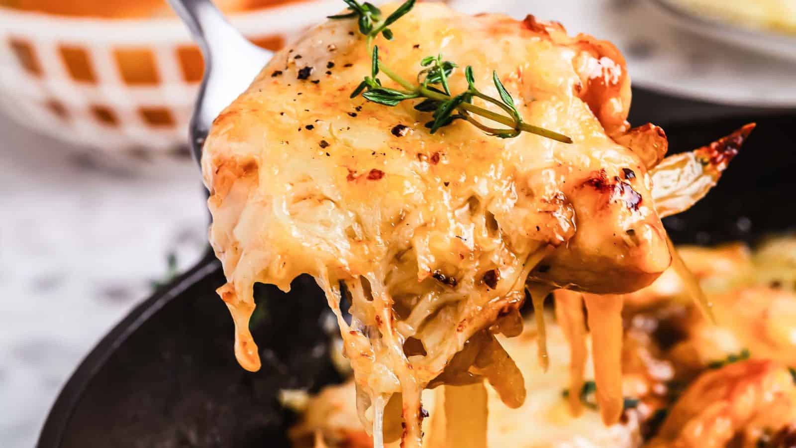 Close-up of a serving of cheesy, baked pasta being lifted with a fork. The dish is topped with melted cheese and garnished with fresh herbs. The background shows a partially blurred bowl.