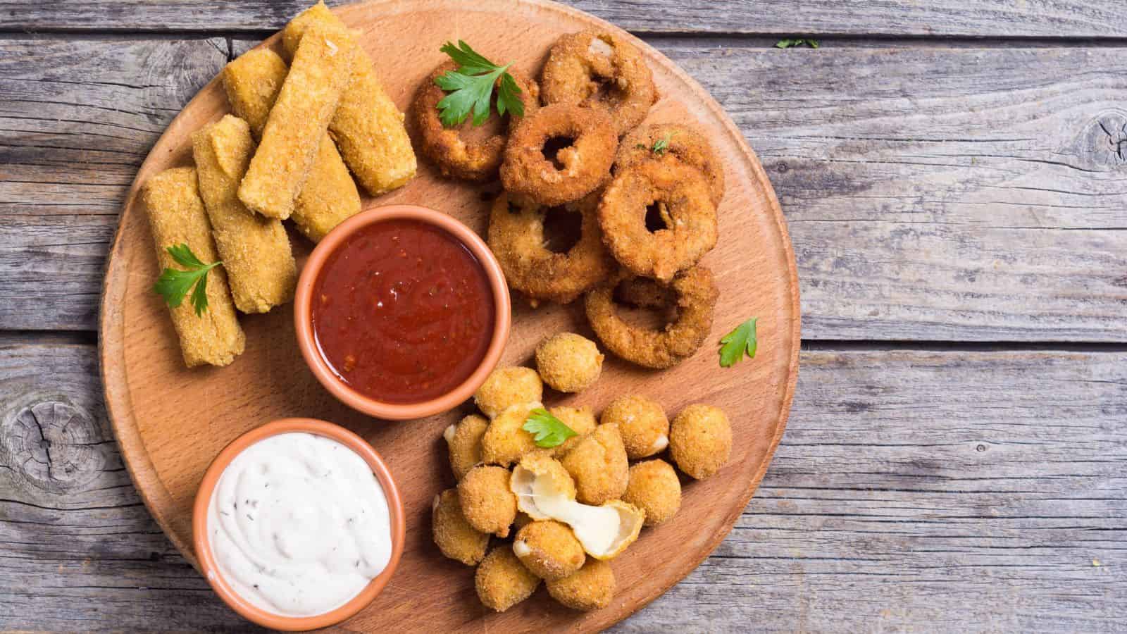 An image of various fried food such as mozzarella bites, calamari, and tofu sticks, there are also two dips, one tomato-based and one white sauce, they are all in a circular wooden board.