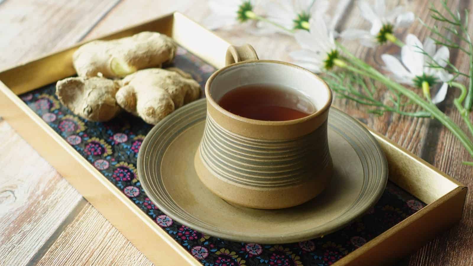 A cup of tea in a ceramic cup and saucer sits on a decorative tray. Fresh ginger roots are placed next to the cup. In the background, white flowers and greenery are partially visible on a wooden surface.