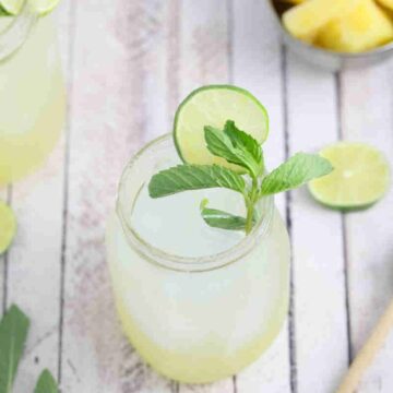 A refreshing drink in a glass garnished with a lime slice and mint leaves sits beside additional lime slices, a honey dipper, and a small bowl of pineapple pieces on the wooden surface. Amidst this delightful setup, an auto draft of warmth flows through the striped cloth in the background.