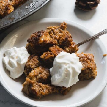 A plate of bread pudding topped with two dollops of whipped cream next to a baking dish with more pudding. A spoon rests on the plate. Nearby are two cinnamon sticks and a small pine cone, all on a light-colored surface.