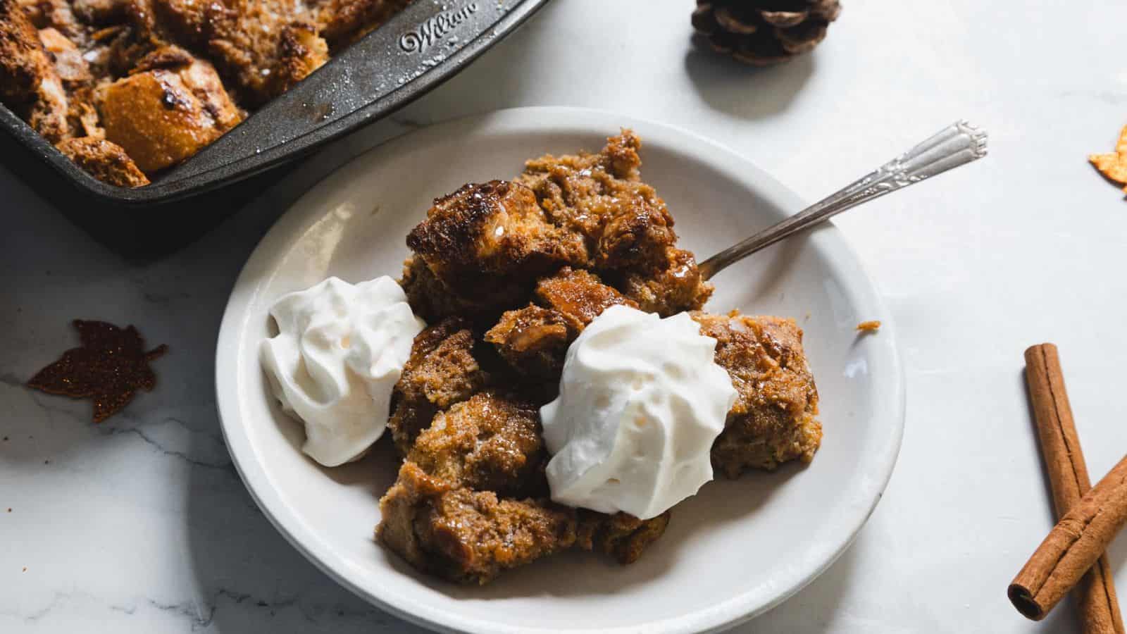 A plate of bread pudding topped with two dollops of whipped cream. A fork rests on the plate. In the background, a baking dish with more bread pudding is partially visible on a marble surface. Decorative items are placed around.