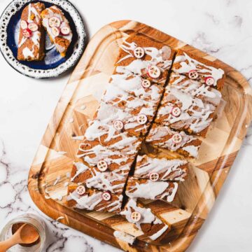 A wooden board holds sliced pumpkin spice cake topped with white glaze and small pumpkin-shaped decorations. A plate beside it contains two additional pieces. A jar and wooden spoon are partially visible on the marble surface, adding a rustic touch to this delightful gluten-free creation.