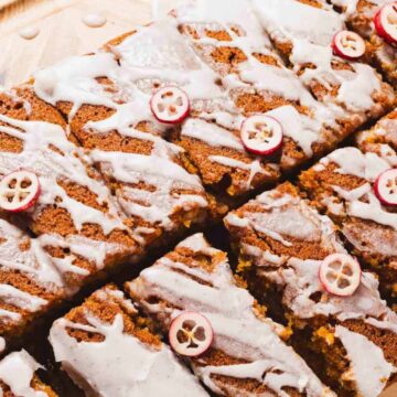 Sliced loaf cake topped with white icing and thin slices of cranberries, arranged on a wooden surface. The cake is cut into rectangular pieces.