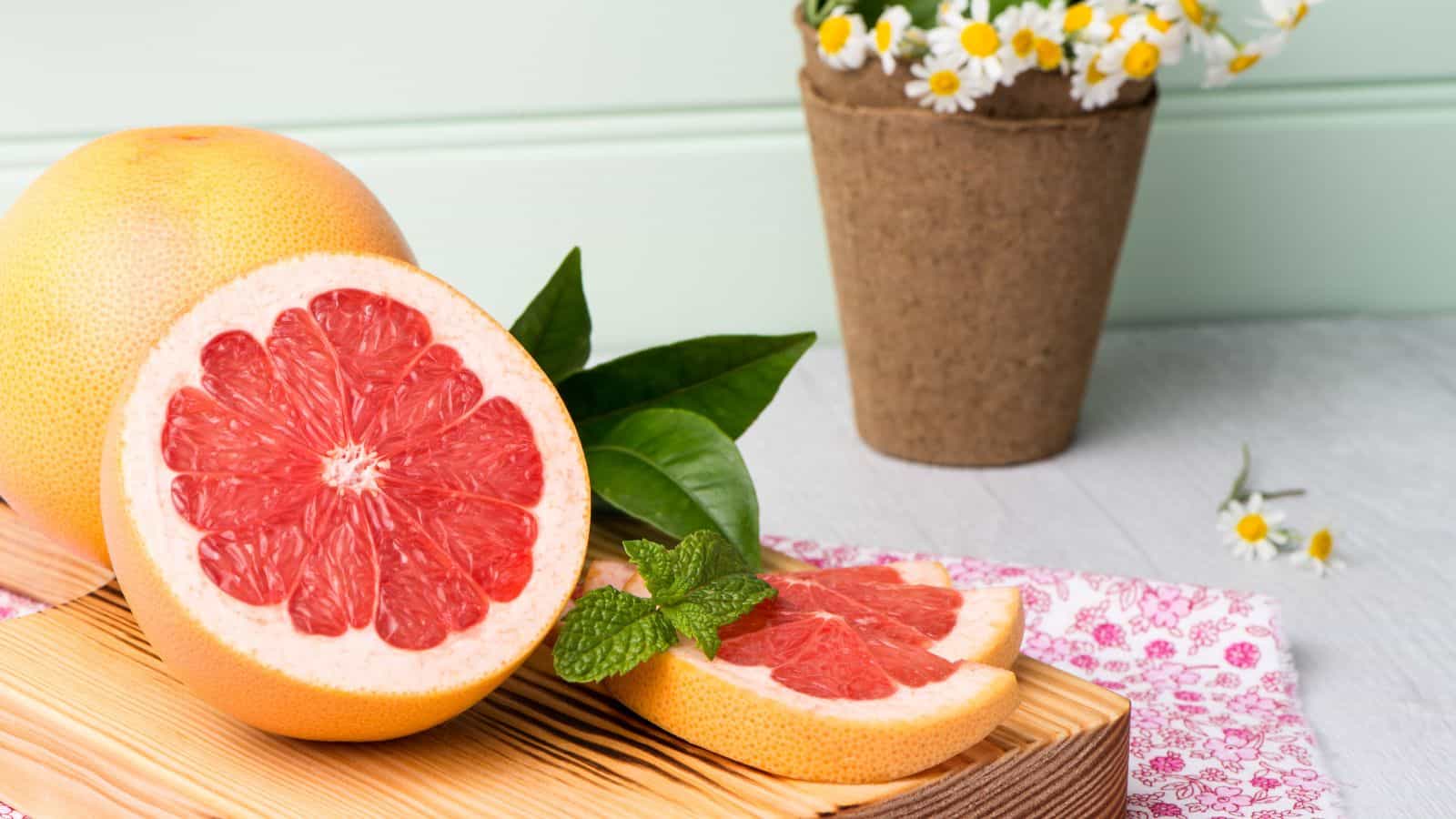 A halved grapefruit and several slices rest on a wooden board, accompanied by fresh mint leaves. In the background, a whole grapefruit and a small pot of daisies are visible. The scene is set on a light-colored surface with pink patterned fabric.