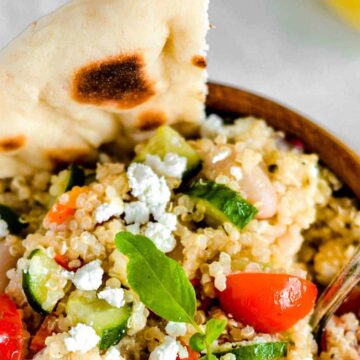 A bowl of quinoa salad, auto-drafted with cherry tomatoes, cucumbers, feta cheese, and fresh greens. A piece of flatbread is on the side, with a spoon resting in the bowl.