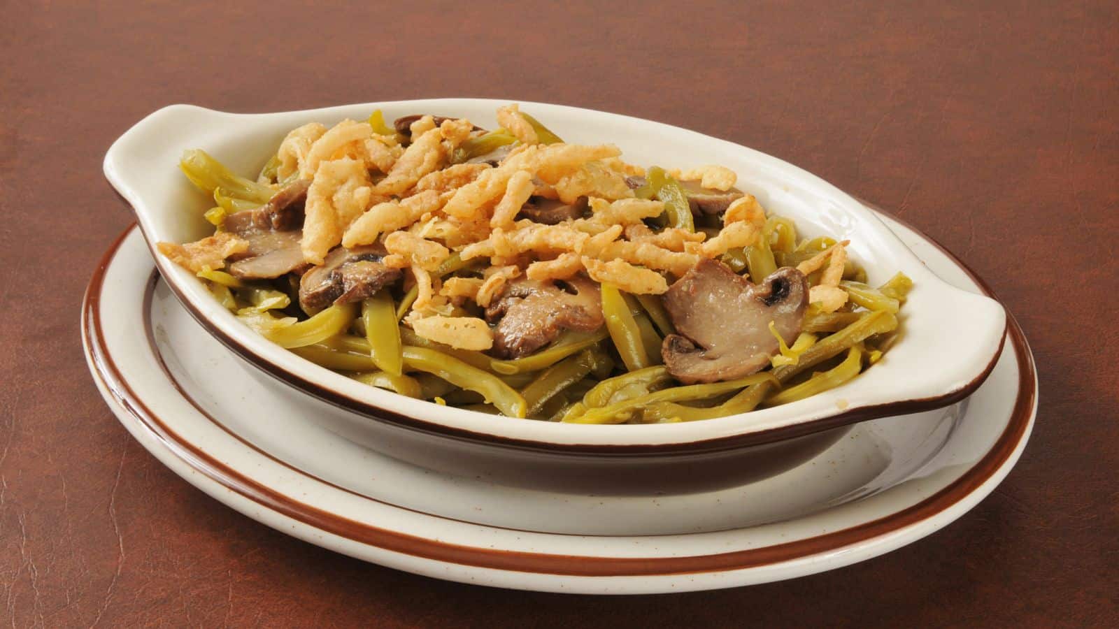 A dish of green bean casserole topped with crispy fried onions and sliced mushrooms is served in an oval ceramic baking dish on stacked plates.