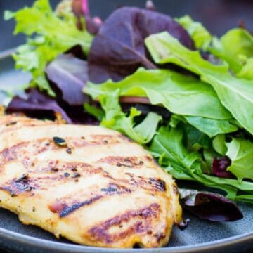 A grilled chicken breast, a star in many grill recipes, is served on a plate alongside a mixed green salad containing various leafy greens. The dish rests on a dark surface, showcasing the chicken's perfect grill marks.