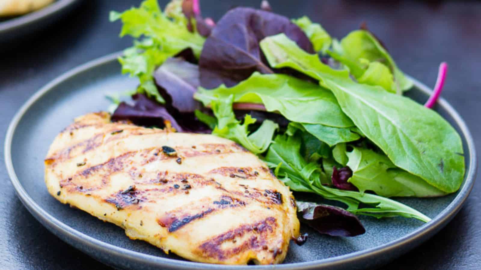 A grilled chicken breast, a star in many grill recipes, is served on a plate alongside a mixed green salad containing various leafy greens. The dish rests on a dark surface, showcasing the chicken's perfect grill marks.