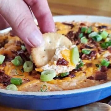A close-up of a dish filled with cheesy dip, topped with chopped green onions and bacon bits—a perfect Christmas appetizer. A hand dips a round cracker into the mixture, all set to wow your guests. The dish rests on a wooden surface with more crackers on the side.