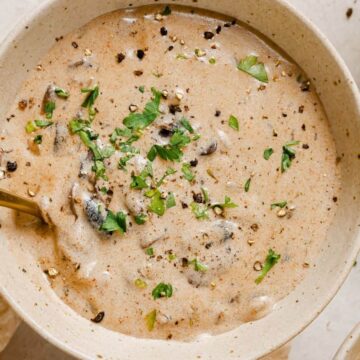 A bowl of creamy mushroom soup, a perfect choice for simple dinners, is garnished with chopped herbs and ground pepper. A spoon rests in the bowl, and the rich, speckled appearance invites you to savor each bite. The bowl sits elegantly on a light-colored surface.