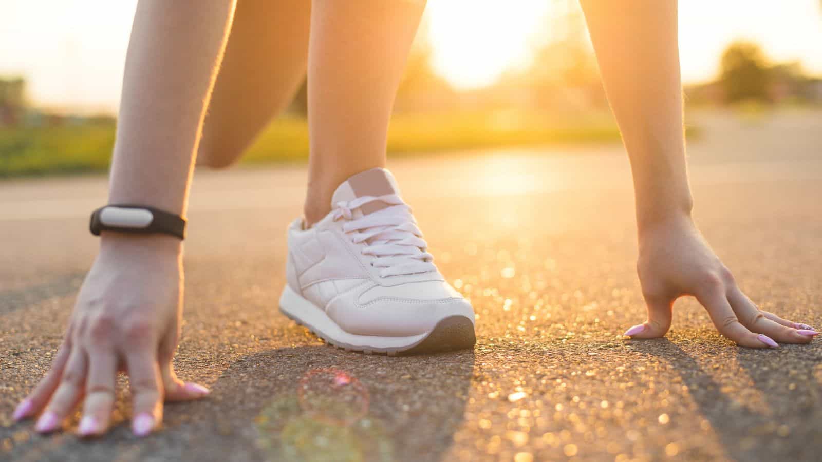 Close-up of a person in a runner's starting position on a paved road, with one hand touching the ground and wearing white athletic shoes. Sunlight casts a warm glow, highlighting a fitness tracker on the wrist—simple tips for balancing holiday feast indulgences.