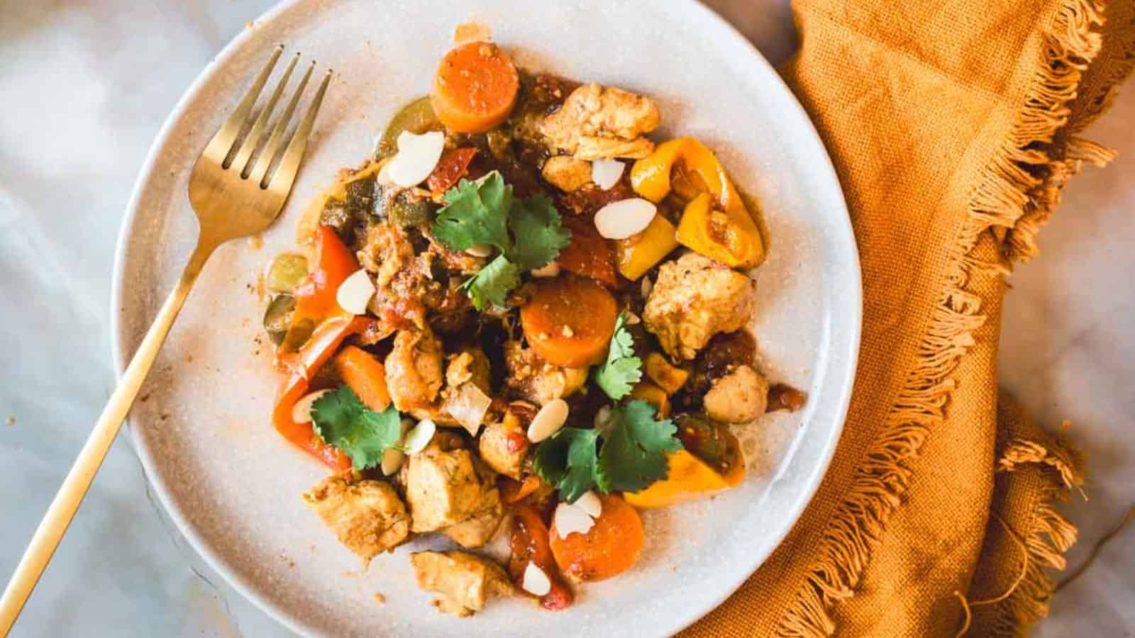 A plate of cooked chicken, sliced carrots, and assorted bell peppers garnished with cilantro and almond slivers. A gold fork is placed beside the plate on a textured orange cloth. The plate is on a marble surface.