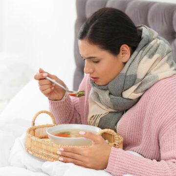 A woman wearing a scarf and a pink sweater is sitting in bed, looking at a spoonful of soup from a bowl she holds on a wicker tray. The background features a gray upholstered headboard and white bedding.