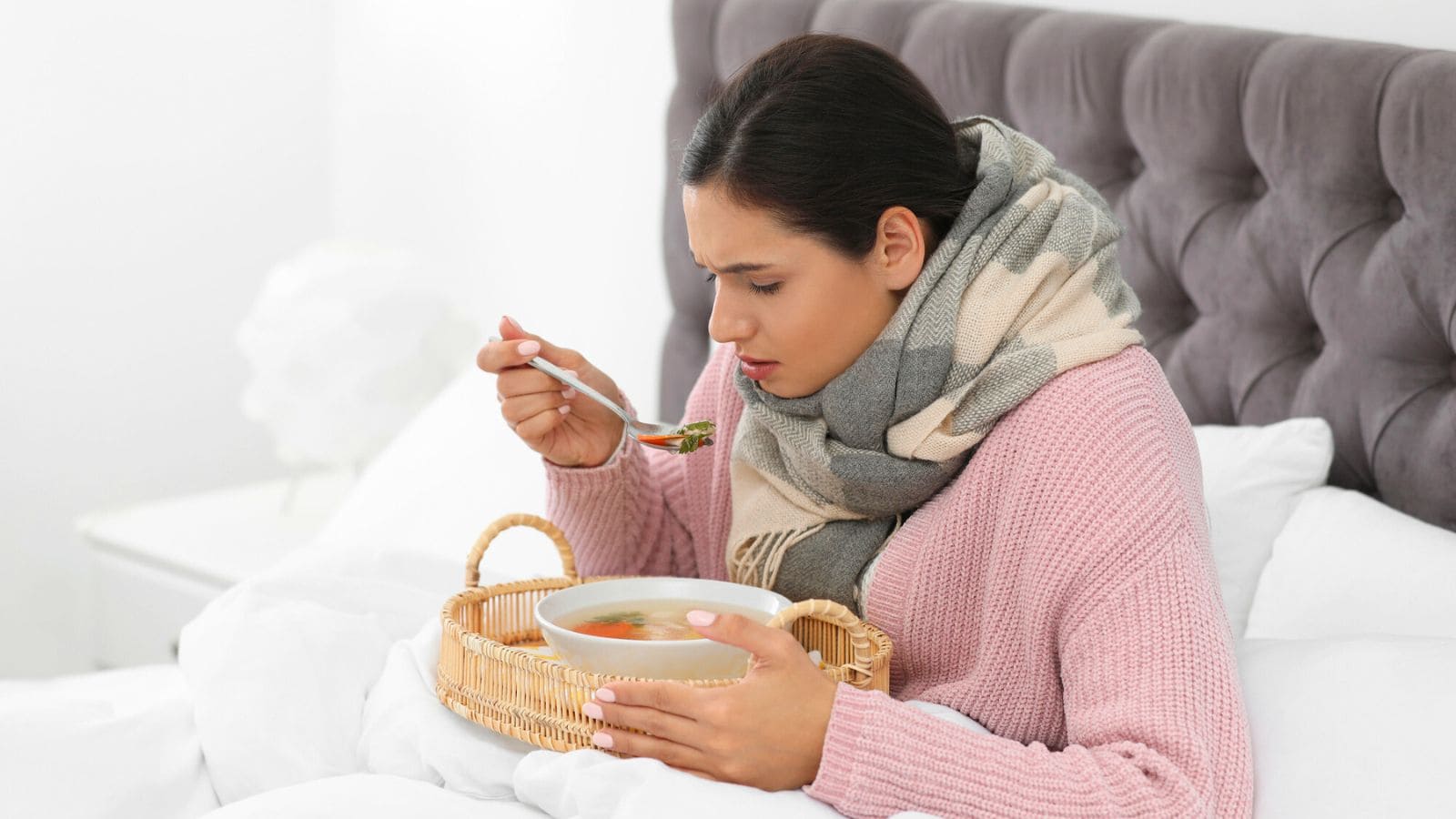 A woman wearing a scarf and a pink sweater is sitting in bed, looking at a spoonful of soup from a bowl she holds on a wicker tray. The background features a gray upholstered headboard and white bedding.
