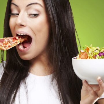 A woman is holding a slice of pizza in her right hand near her mouth and a bowl of salad in her left, indulging in what seems like an innocent bite. The background is a solid green color, playfully hinting at how sneakily high in sugar some foods can be.