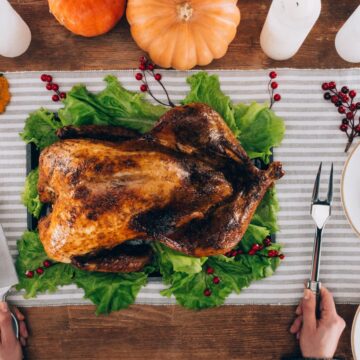 A beautifully roasted Thanksgiving turkey on a bed of lettuce, surrounded by dishes of grilled corn, carrots, and Brussels sprouts on a striped tablecloth, with pumpkins and candles as festive decor.