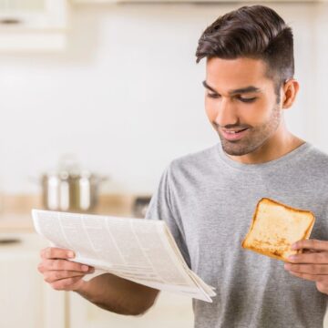 A person stands in a kitchen holding a slice of toast in one hand while reading a newspaper with the other. They wear a gray t-shirt and appear to be in a well-lit, modern kitchen setting. Cooking pots are visible in the background.
