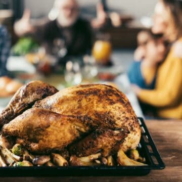 A roasted turkey sits on a tray with vegetables in the foreground. In the blurred background, a family is gathered around a dining table, engaging in conversation.