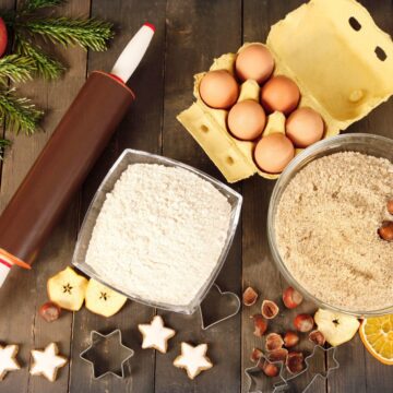 A flat lay of baking ingredients on a wooden surface sets the stage for holiday meal planning tips. Items include a rolling pin, a carton of eggs, a bowl of flour, nuts and oats, whole nuts, apple and orange slices, star-shaped cookie cutters, and a sprig of evergreen.