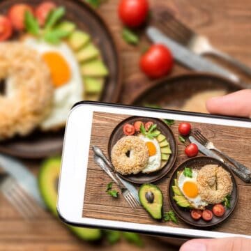 A person holds a smartphone, capturing a photo of a wooden table adorned with a plate featuring an Instagram-worthy bagel, fried egg, avocado slices, and cherry tomatoes. Another similar plate teases in the background, while utensils and mint leaves add to the hype of these drool-worthy foods.