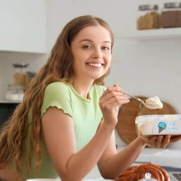 A person with long hair smiles while holding a spoon and a tub of ice cream in a kitchen. They're wearing a green shirt and standing near a countertop with a laptop and a few jars of ingredients in the background.
