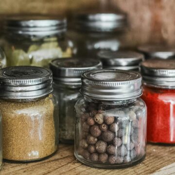 A wooden shelf beautifully displays various glass jars filled with spices and common herbs. The jars, topped with metal lids, are arranged in neat rows, showcasing the vibrant colors and textures of red, green, brown, and white—each offering its unique health benefits.