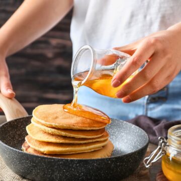A person pours syrup from a glass container onto a stack of pancakes in a frying pan. A jar of syrup and honeycomb are nearby on a wooden surface.