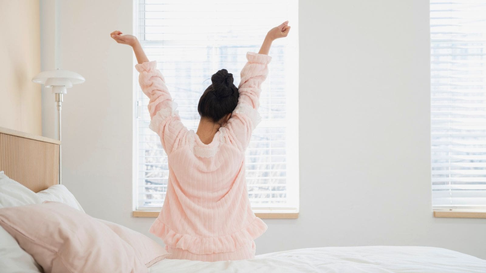 A person wearing pink pajamas is sitting on a bed and stretching with arms raised. The room is softly lit by sunlight coming through the blinds of two windows. A lamp is visible beside the bed.