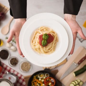 A person holds a plate of spaghetti topped with tomato sauce and basil, embodying a slice of Italian culture. Below, the table brims with vegetables, herbs, and seasonings, inviting American visitors to explore traditional Italian eating habits in this vibrant cooking setting.