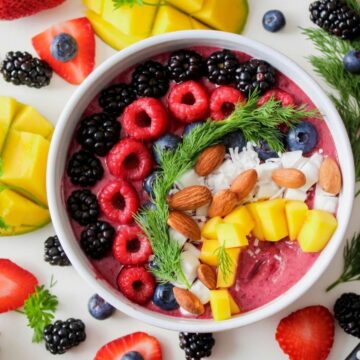 A colorful smoothie bowl topped with raspberries, blackberries, almonds, coconut flakes, and mango pieces, garnished with sprigs of dill. These surprising superfoods are complemented by fresh strawberries and sliced mango surrounding the bowl on the table. A spoon is nearby.