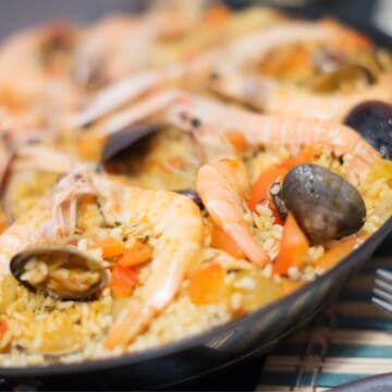 A close-up of a seafood paella in a black pan showcases shrimp, mussels, clams, and diced vegetables nestled in rice. This dish reminds one of regional dishes in the US with interesting stories. A fork rests on the table beside the pan, set atop a striped placemat.