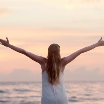 A person with long hair is standing on a beach at sunset, facing the ocean with arms outstretched. The sky is a blend of soft pink and orange hues, creating a serene and peaceful atmosphere.