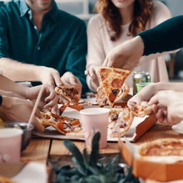 A group of people seated around a wooden table are reaching for slices of pizza from various pizza boxes, enjoying a mix of traditional dishes. Drinks and other food items are scattered on the table. A plant is in the foreground, slightly out of focus.
