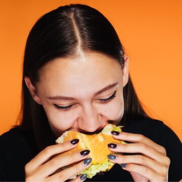 A girl biting into a burger in front of an orange background.