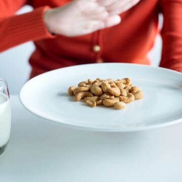 A person wearing an orange sweater is cautiously pushing away a plate of peanuts with one hand to avoid allergies. A glass of milk sits on the table beside the plate, emphasizing their careful choice of foods to prevent a flare-up.